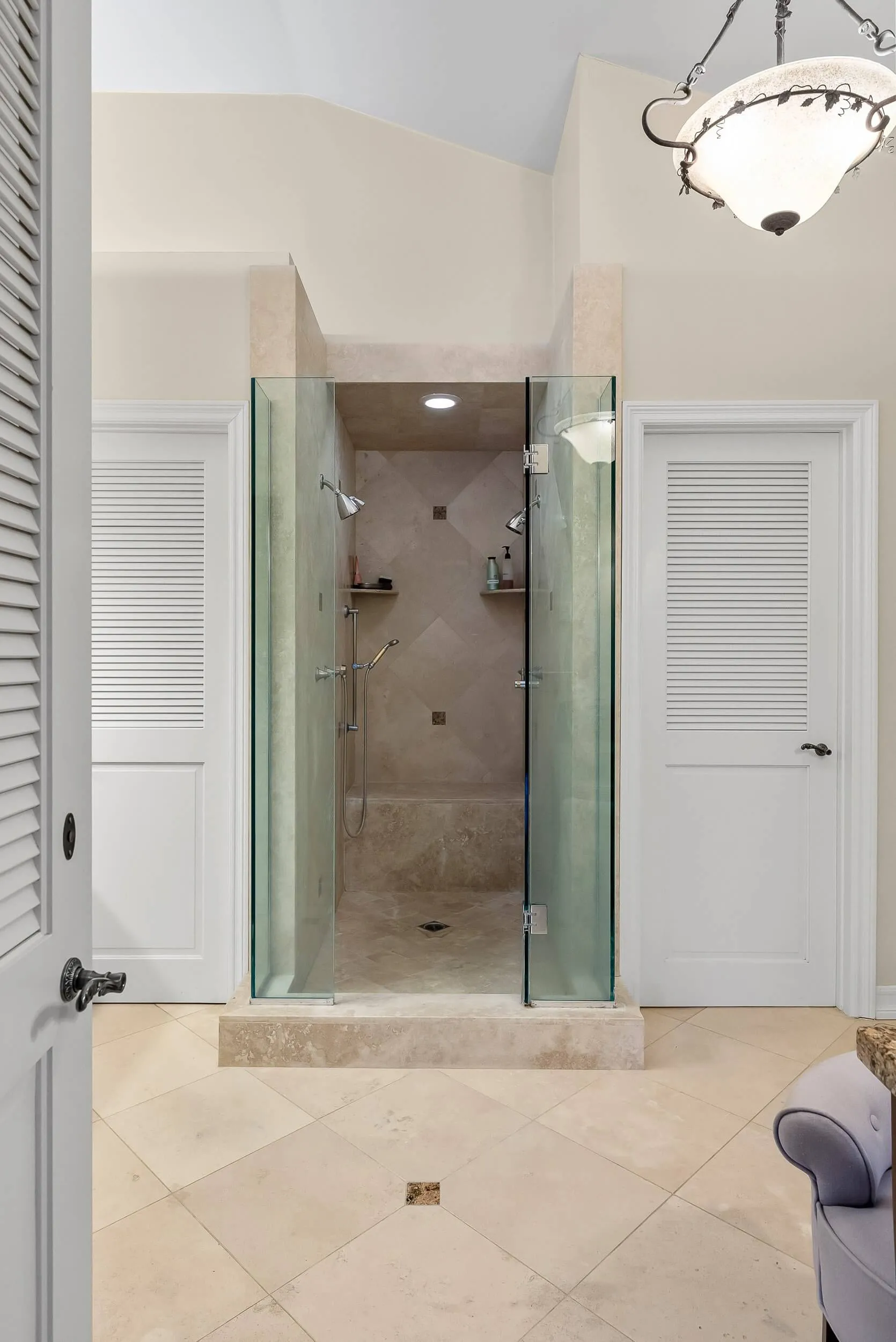Walk-in glass shower with travertine tile flanked by louvered storage doors