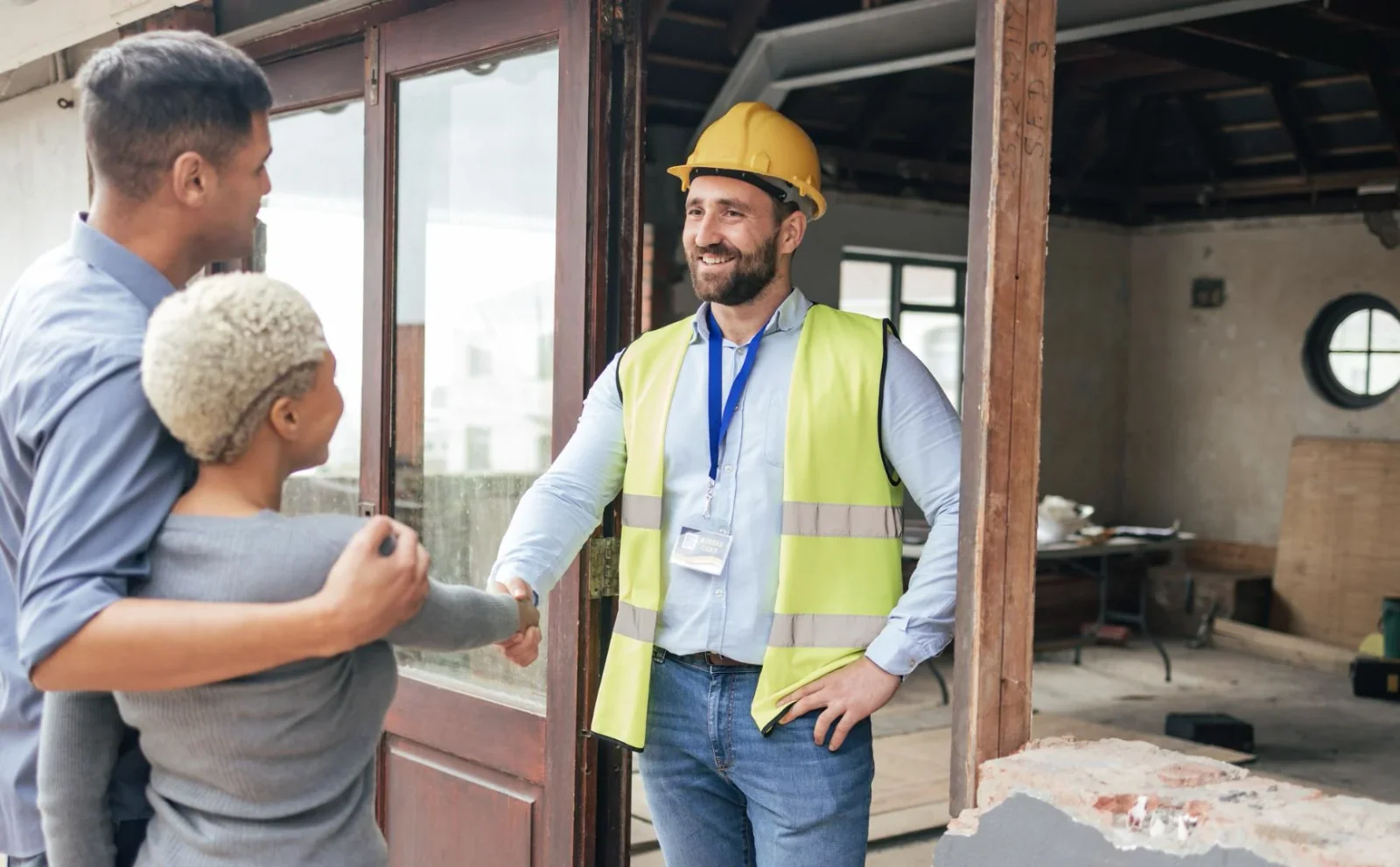 Contractor in a safety vest shaking hands with clients inside a building under renovation