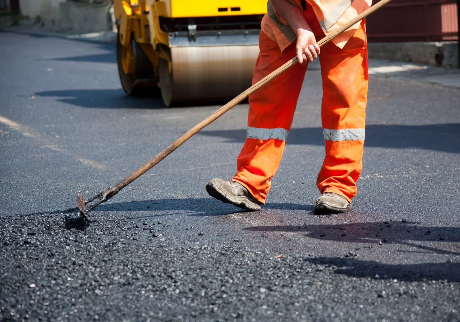 Worker in orange spreading fresh asphalt with a road roller behind him
