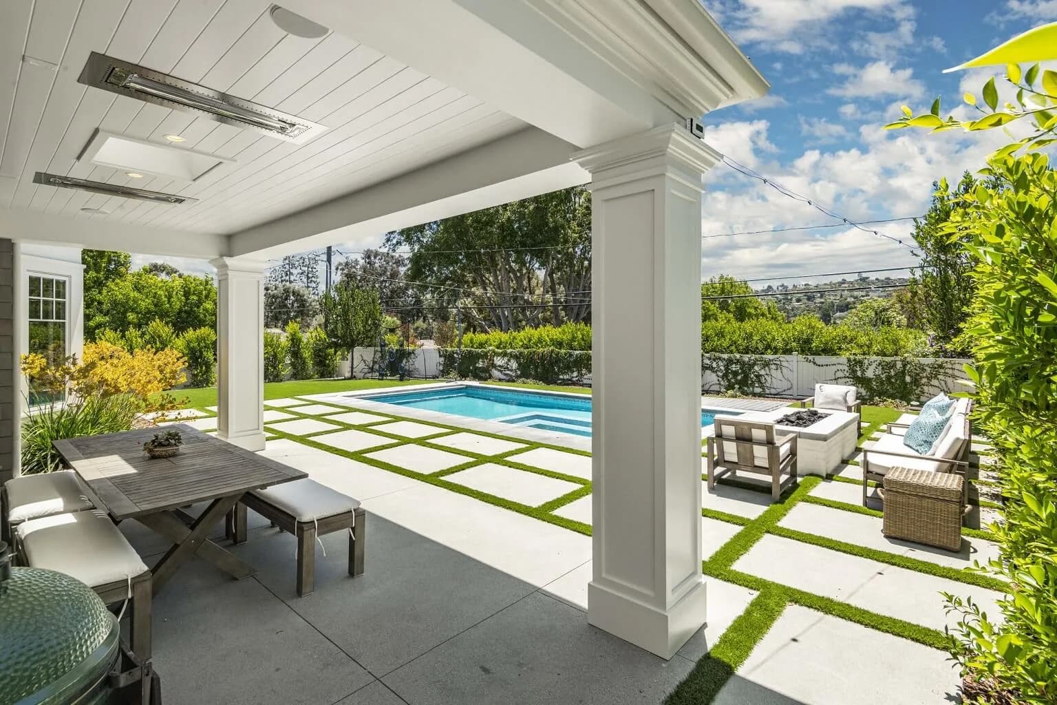 Covered outdoor patio with dining table and overhead heaters overlooking the pool and lawn